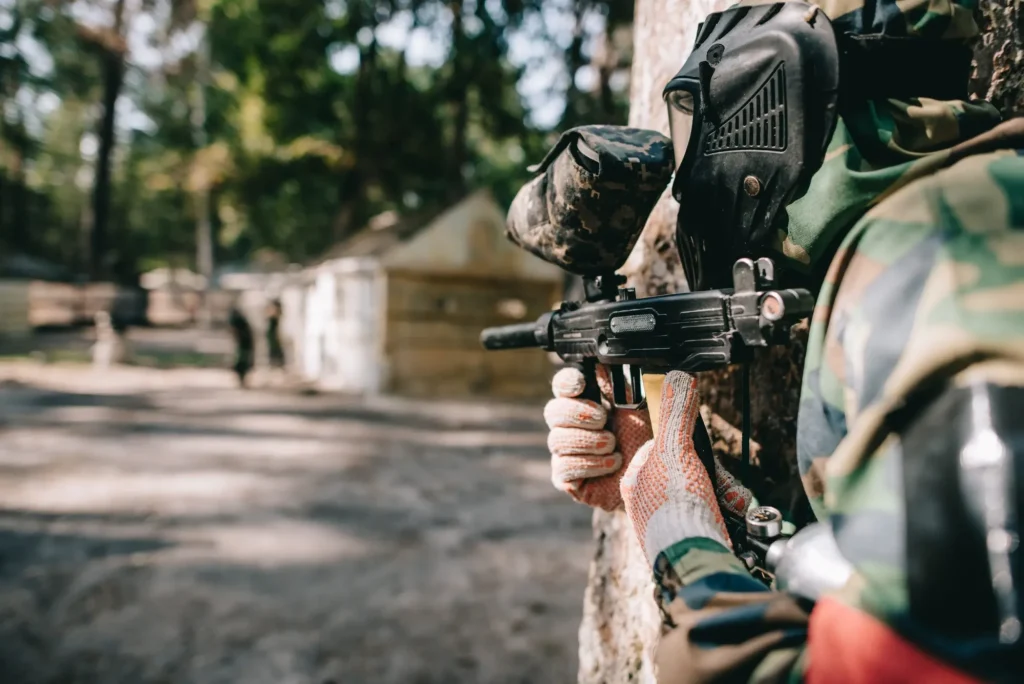 selective focus of paintball player in goggle mask and camouflage aiming by paintball gun outdoors