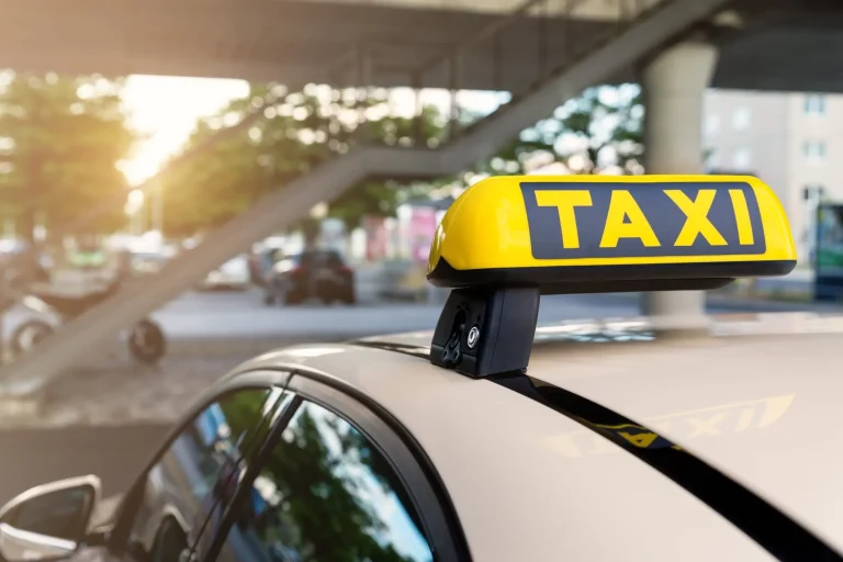 Close-up detail yellow taxi symbol on cars roof stand waiting at parking of airport terminal or railway station against park warm evening bokeh sunlight. Urban street transportation comfort service